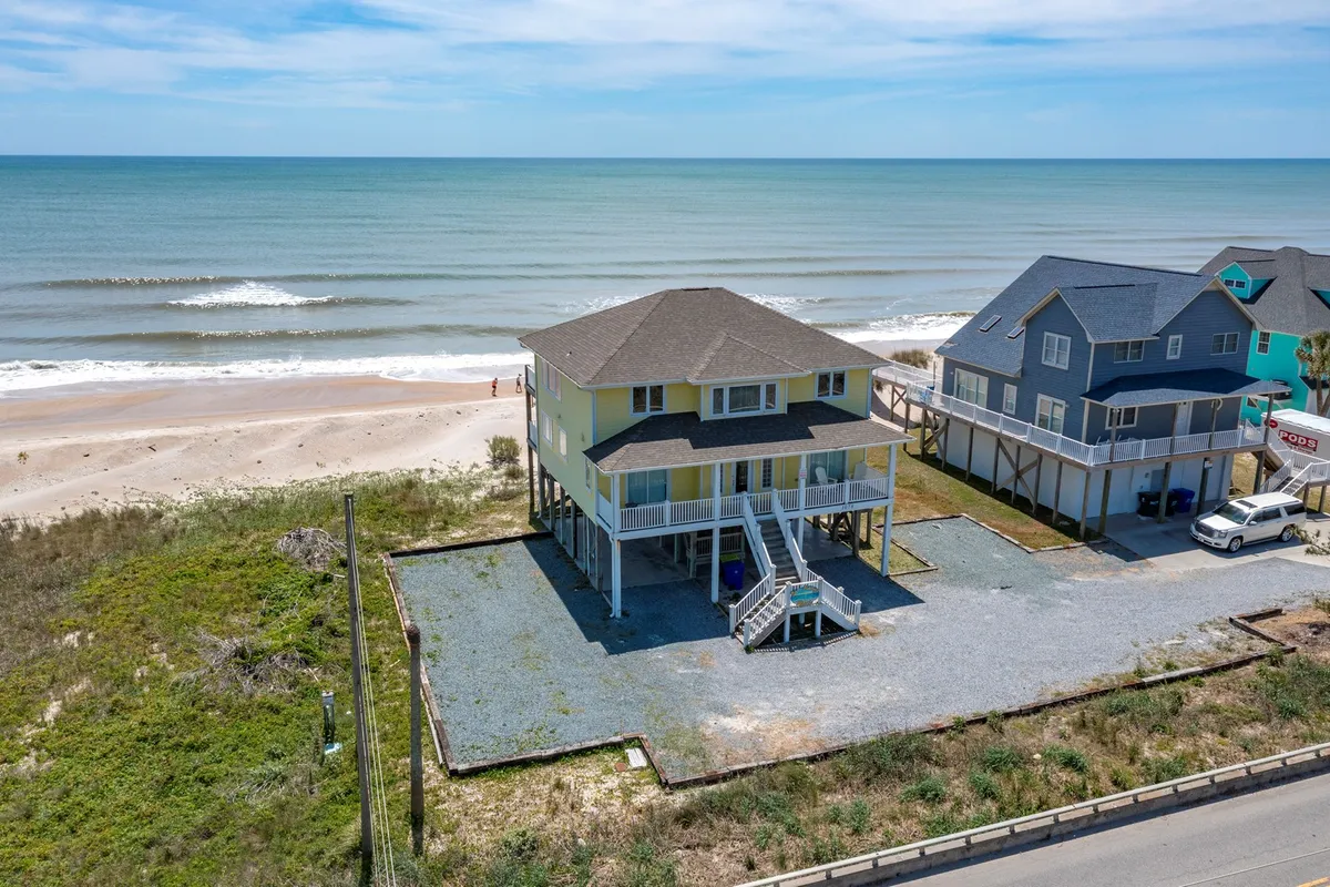 A peaceful beachfront house stands just beyond gentle sand dunes, with rolling ocean waves stretching toward the horizon. Neighboring properties are visible, hinting at the welcoming community feel. The scene evokes early mornings, with soft light ideal for sunrise watching or setting out for beachcombing and outdoor adventure.