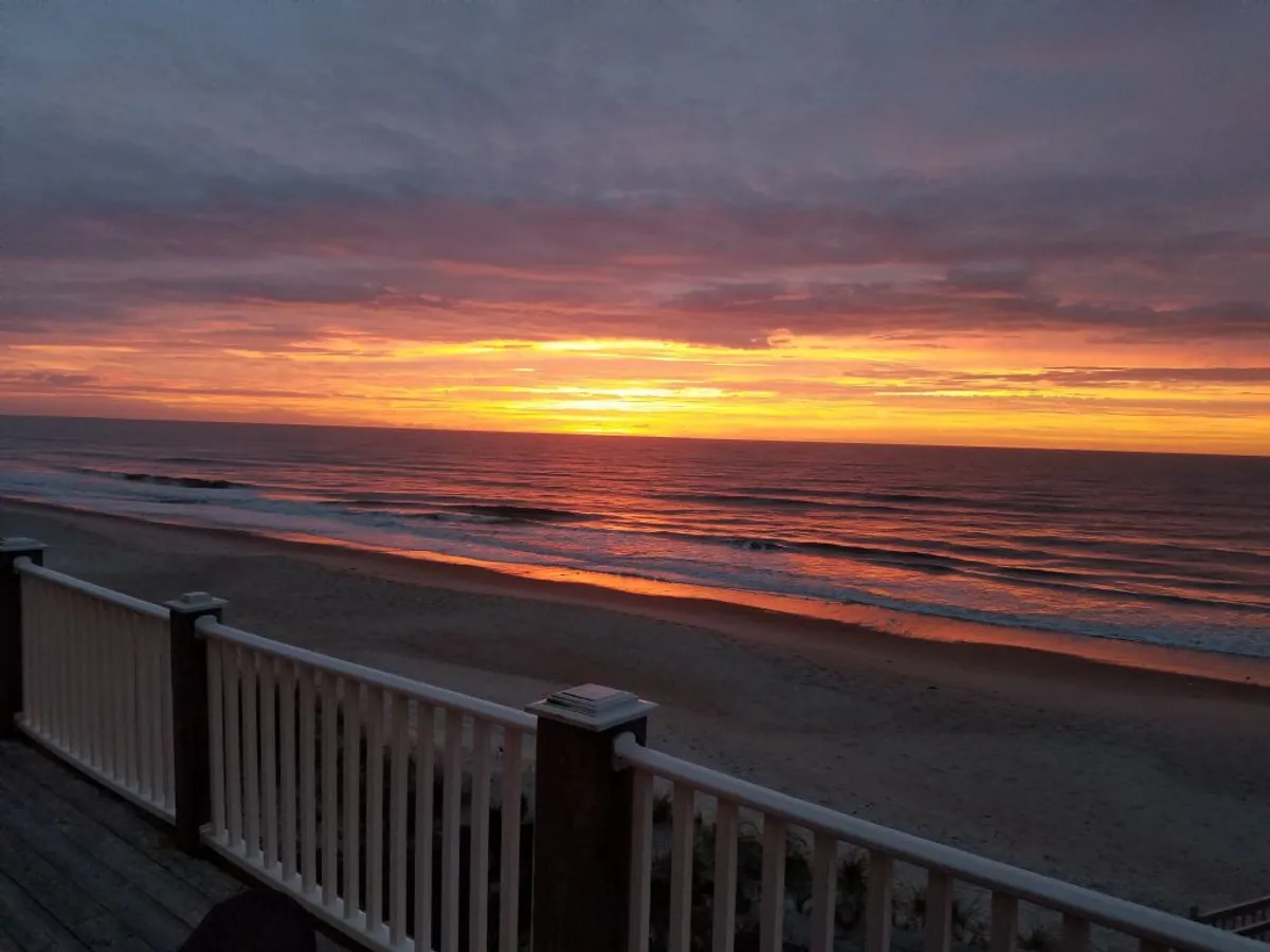 A tranquil sunset scene from a deck or balcony, with the ocean waves and sandy beach visible beyond a sturdy railing. The soft evening light offers an inviting atmosphere for reflection, connection, or group gatherings at the end of the day.
