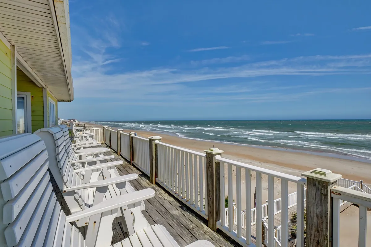 A tranquil deck scene featuring white wooden chairs positioned to face a stunning ocean view, with the beach shore just beyond the rail. The setup invites relaxation, sunbathing, reading, or watching the sunrise and sunset with loved ones.