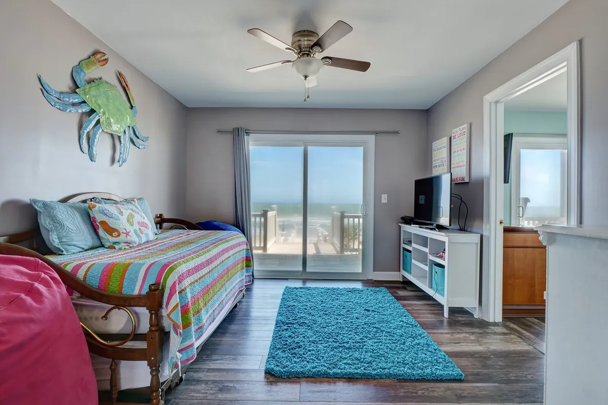 A bright, playful bedroom with a colorful bedspread, large crab wall art, sliding glass doors leading to a balcony with ocean views, and natural light filling the room. The space exudes a cheerful, family-friendly vibe.