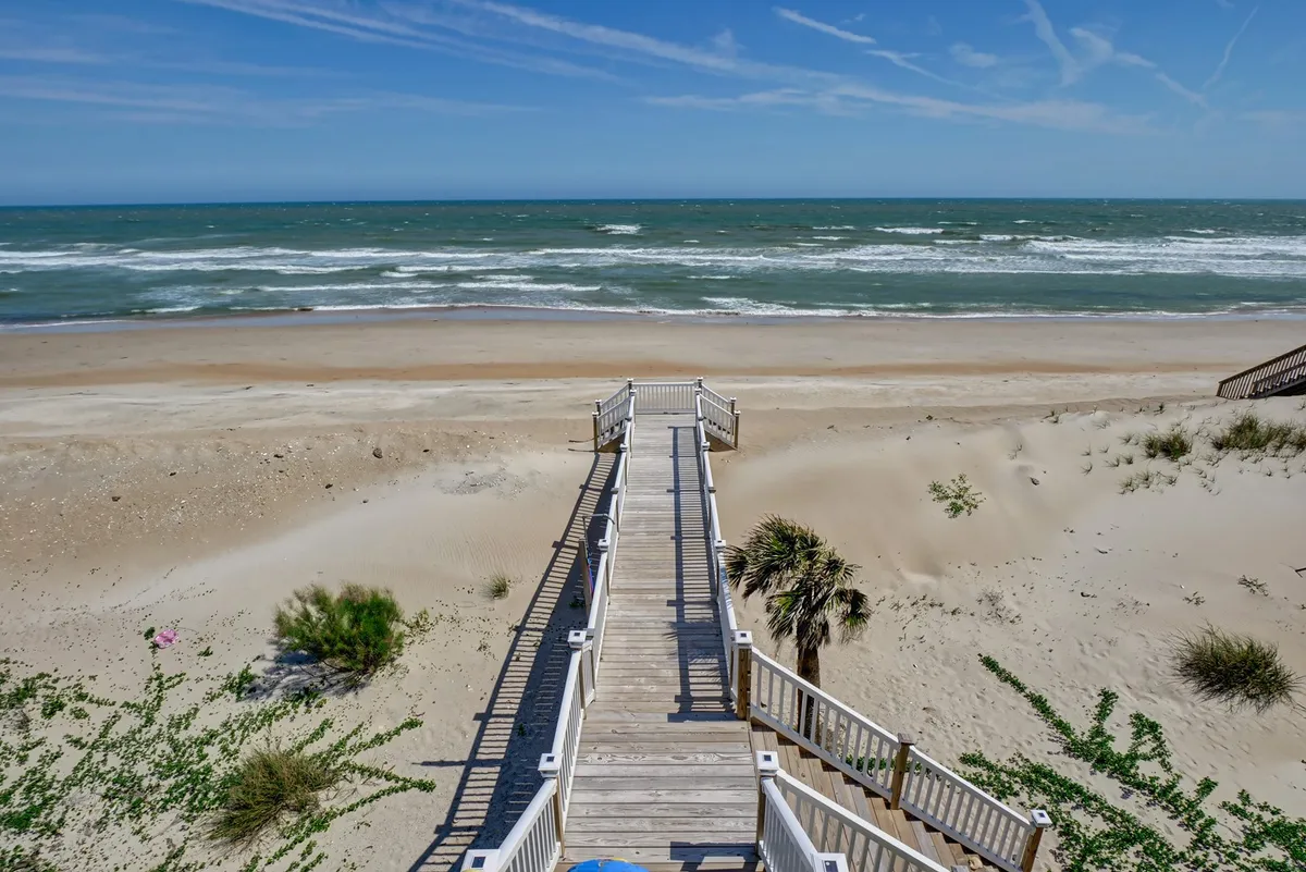 This image features a wooden walkway stretching out toward the sandy beach, with gentle ocean waves rolling in and a distant horizon illuminated by soft morning light. The scene captures the tranquil, direct connection from the property to the beach, with hints of people walking, sunbathing, and exploring the shore.