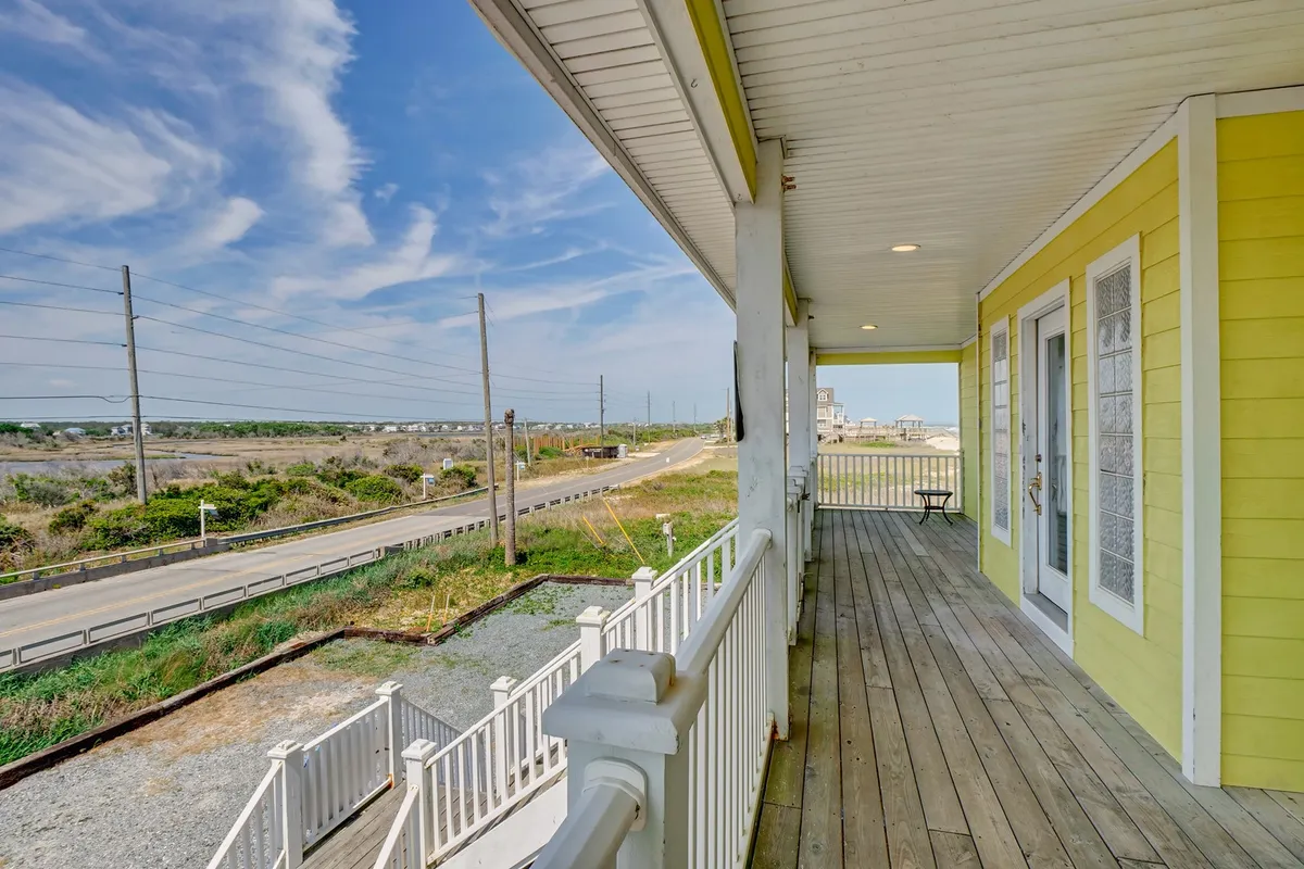 The exterior balcony of the yellow house, featuring a clear railing and open views to the surrounding landscape. The scene captures a peaceful moment with potential for morning coffee, group chats, or enjoying the coastal breeze as a group.
