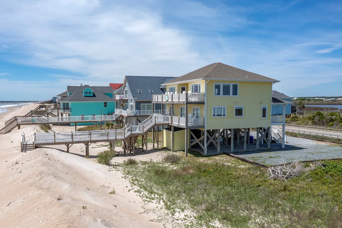 A beachfront house stands proudly beyond a boardwalk, surrounded by other coastal homes. The inviting scene includes the promise of gatherings, with the ocean as a constant, calming backdrop. The deck is visible, hinting at relaxing family meals with ocean views.