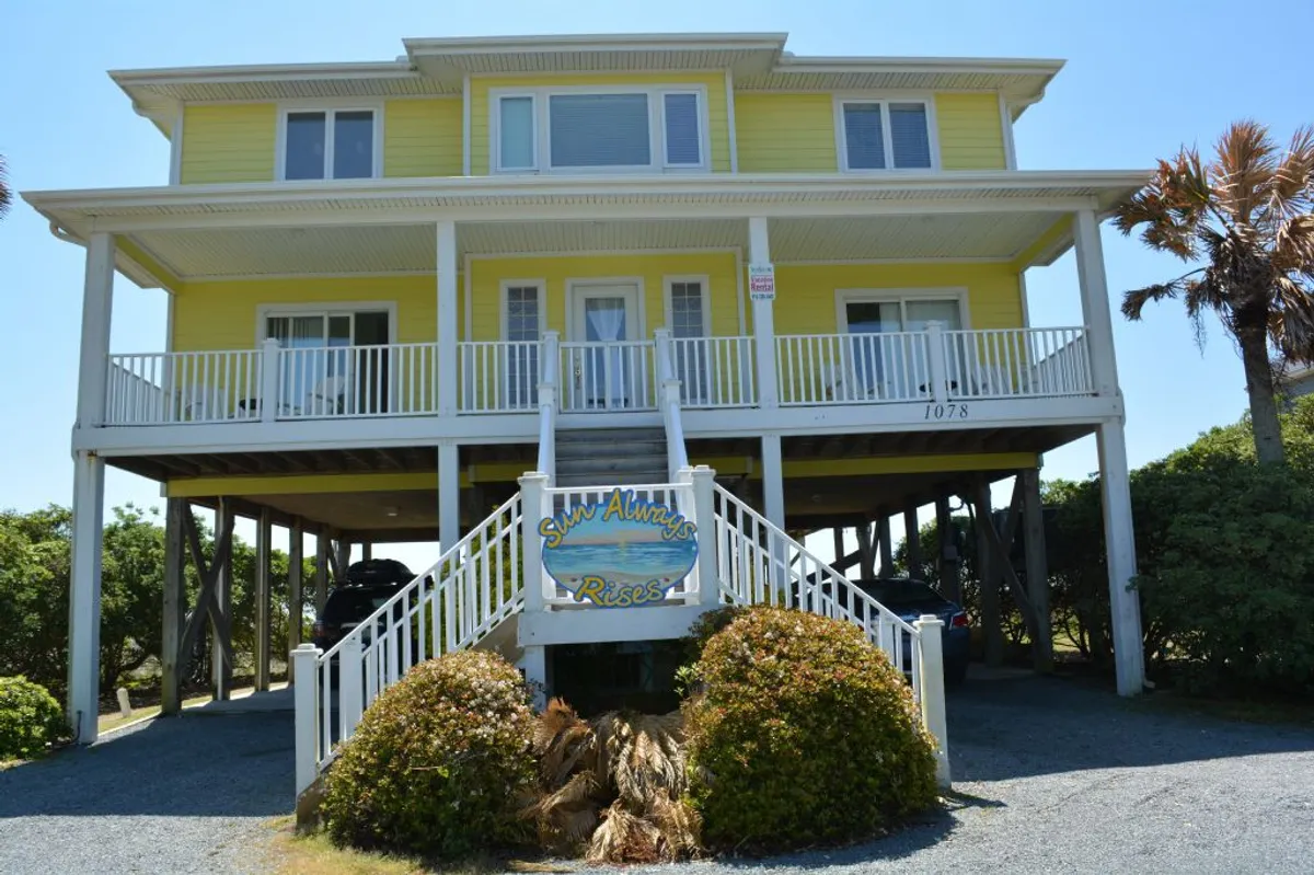 The cheerful yellow exterior of 'Sun Always Rises,' framed by lush palm trees and a decorative sign, welcomes guests and their pets. The inviting steps and porch hint at restful moments shared outdoors, perhaps with a dog relaxing nearby.