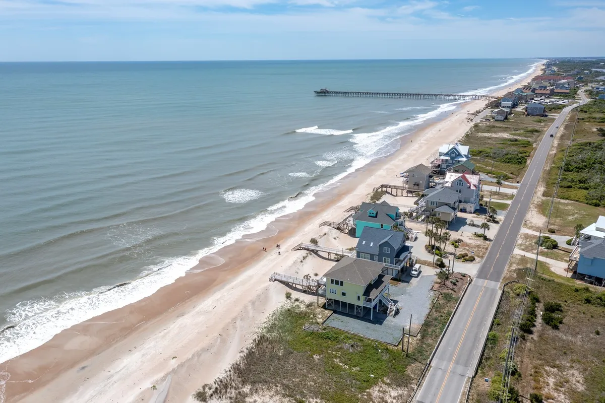 A wide stretch of sandy beach meets the rolling ocean, with a scenic pier stretching out over the water. Seaside houses line the shore, and a quiet coastal road winds behind them. The relaxed mood is perfect for imagining sunrise walks, fishing, swimming, or simply enjoying the fresh air.