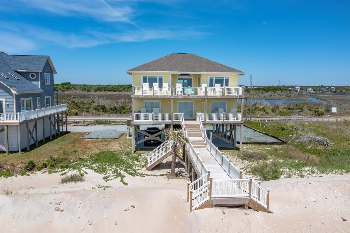 The image showcases a charming beachfront single-family house in North Topsail Beach, North Carolina. A private wooden walkway leads from the spacious deck through lush greenery directly to the sandy shoreline. The ocean is visible in the background, with soft sunset lighting casting a warm, inviting glow over the property, evoking a sense of peace and relaxation.