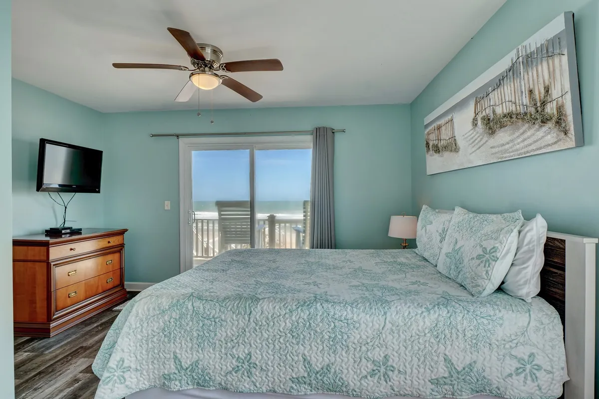 A tranquil bedroom featuring a king-sized bed with aqua-colored bedding, a large window that offers an expansive ocean view, a wooden dresser, and a wall-mounted television. The decor is serene, with soft natural light enhancing the restful atmosphere.