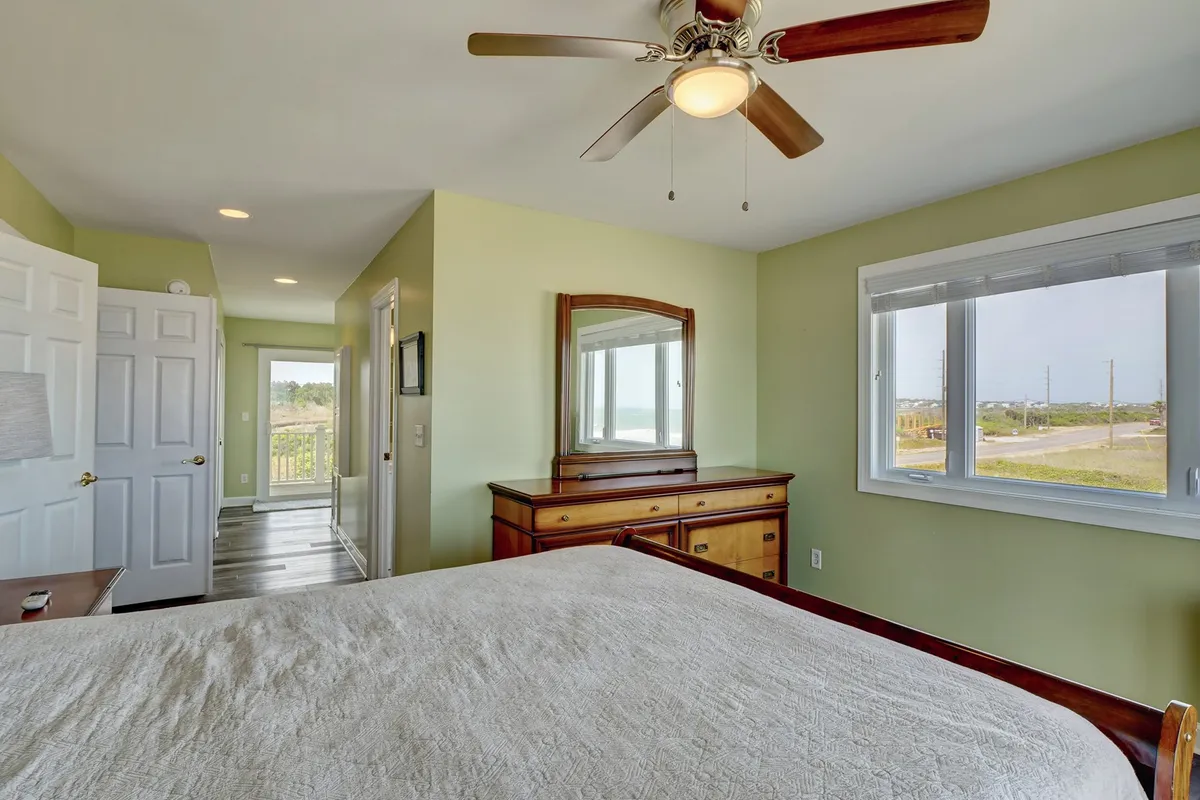 A serene bedroom with light-colored bedding, wooden dresser, and windows offering a peaceful view of nature. The mirror reflects natural light throughout the room, enhancing the soothing, tranquil ambiance.