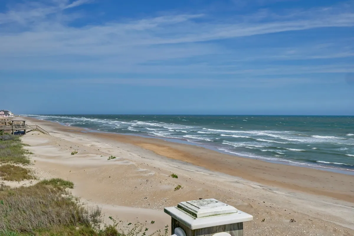 A wide, panoramic view of the vast ocean, golden sand, and rolling dunes under a brilliant blue sky. The open, family-friendly beach is dotted with possibilities for building sandcastles, picnicking, and enjoying serene beach days.