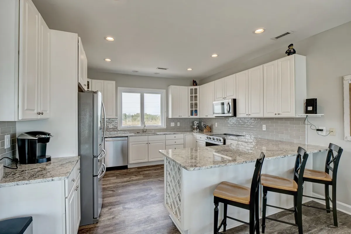 A bright and welcoming kitchen featuring a granite countertop, stainless steel appliances, a spacious kitchen island with seating, and large windows that fill the space with natural light. The layout is open and ideal for preparing meals or sharing morning coffee.