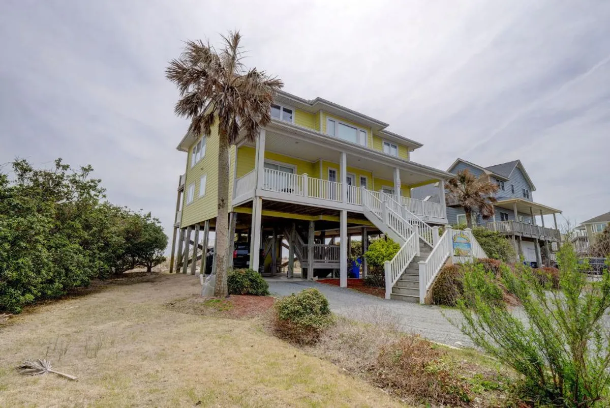 A bright yellow beach house surrounded by lush palm trees, with wide, sturdy stairs leading up to a spacious porch. The home is set against a coastal backdrop, suggesting proximity to ocean views and outdoor gatherings.
