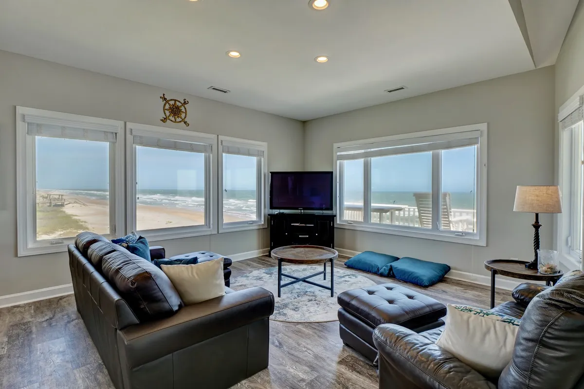 A comfortable living room with cozy seating, a coffee table, and a large television, all set against a backdrop of large windows offering ocean views. The space feels tranquil and inviting, perfect for gathering in the evenings.