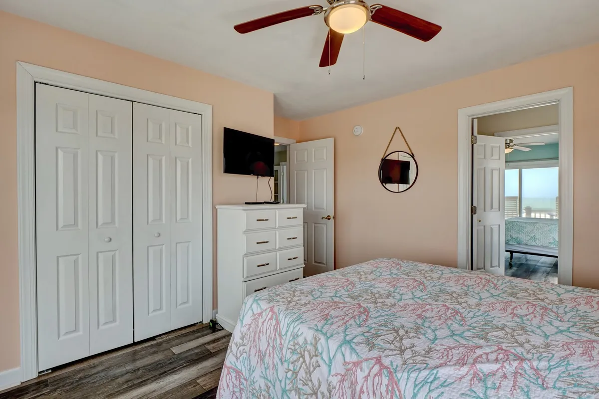 A serene bedroom featuring a bed with coral-patterned bedding, wall-mounted TV, ample dresser space, and a ceiling fan. Gentle sunlight fills the room, creating a cozy environment ideal for rest and quiet moments.