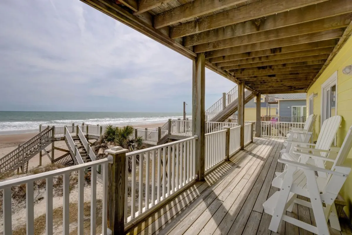 A peaceful deck area with comfortable seating, stairs leading directly down to the sandy beach, and the shoreline visible in the background. The image evokes a sense of immediate access to the beach for guests and pets alike, with a seamless flow from deck to sand.